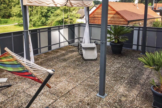 La terraza en la azotea con antiguas baldosas de hormigón ofrece una amplia zona de estancia. La superficie sucia de las baldosas muestra claros signos de desgaste. El toldo proporciona una agradable sombra en los días calurosos. Las macetas con palmeras y una hamaca crean un ambiente acogedor. La barandilla de metal y vidrio completa el diseño moderno y abierto de la terraza.