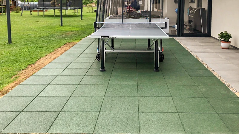 En una terraza con baldosas verdes WARCO, elaboradas con granulado de caucho aglutinado con PU, se ha colocado una mesa de ping-pong frente a una casa lujosa. En el jardín hay un trampolín y otros juegos que invitan al entretenimiento activo y al ejercicio. Las baldosas robustas y antideslizantes brindan comodidad y seguridad. Su superficie elástica protege las articulaciones, mientras que su diseño de fácil mantenimiento mantiene un aspecto impecable.