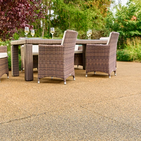 Terraza soleada en un jardín con losas WARCO beige-marrón de granulado de caucho ligado con PU. La superficie antideslizante y permeable al agua ofrece una pisada cómoda y reduce el ruido. Una mesa de jardín robusta con sillas trenzadas y copas de vino invita a relajarse. Arbustos frondosos y flores de colores generan un ambiente natural. Este pavimento elástico es resistente a las heladas, mantiene su forma y resulta ideal para todo el año.