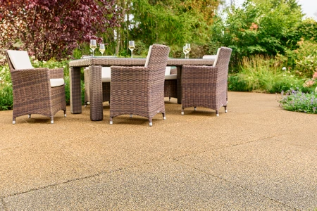 Terraza soleada en un jardín con losas WARCO beige-marrón de granulado de caucho ligado con PU. La superficie antideslizante y permeable al agua ofrece una pisada cómoda y reduce el ruido. Una mesa de jardín robusta con sillas trenzadas y copas de vino invita a relajarse. Arbustos frondosos y flores de colores generan un ambiente natural. Este pavimento elástico es resistente a las heladas, mantiene su forma y resulta ideal para todo el año.