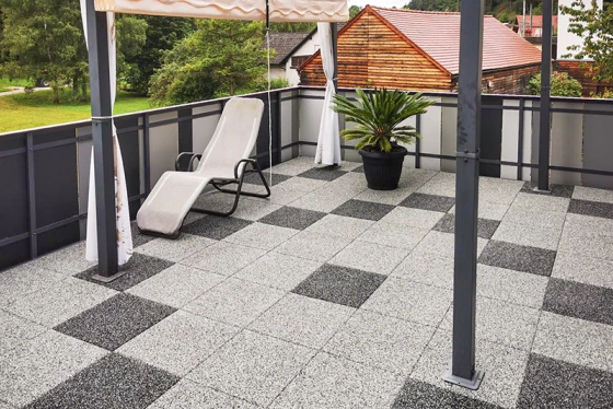 La terraza de azotea renovada cuenta con baldosas efecto granito dispuestas en un patrón geométrico de cuadrados grises claros y oscuros, creando un ambiente moderno y elegante. El pavimento de baldosas de goma robusto proporciona seguridad antideslizante y comodidad. Un toldo ofrece sombra agradable en los días calurosos. Una maceta con palmera y una tumbona completan el diseño acogedor de la terraza. La barandilla de metal y vidrio aporta un aspecto abierto y aireado.