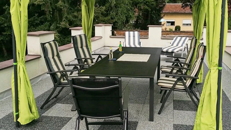 En una terraza en la azotea, un suelo compuesto por baldosas de terraza WARCO luce un efecto jaspeado en tonos gris claro y gris oscuro, evocando el granito. Un baldaquín protege la zona de asientos sobre esta superficie elástica y resistente a la intemperie. Las baldosas antideslizantes y amortiguadoras se instalan sin adhesión permanente y reducen el ruido de pisadas. El resultado es un espacio de fácil mantenimiento, perfecto para relajarse al aire libre.