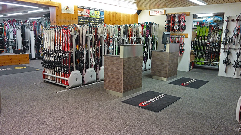Behind two counters in the Gaby Sports ski shop are stands with various ski models. The floor is covered with indoor click tiles from WARCO in the colour Rattan Lounge.