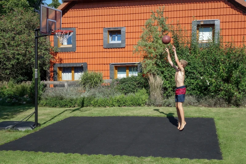 Ein Junge wirft den Basketball auf den Basketballkorb. Den kleinen Basketballplatz aus schwarzen Ballspielplatten von WARCO hat die Familie selbst im Garten angelegt.
