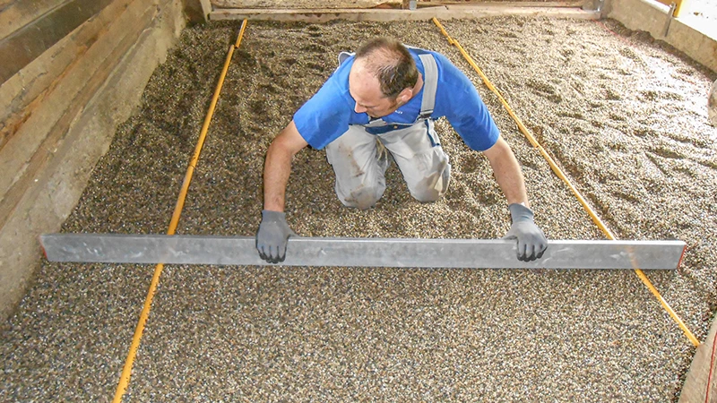 Instalación de un nuevo suelo en una cuadra para caballos: Un hombre nivela una capa de grava con una regla. La grava sirve de base para colocar un suelo de goma granulado en los boxes. La zona está delimitada lateralmente por paredes de madera o de hormigón. El trabajo se realiza con cuidado y precisión, como corresponde a una base de cuadra bien ejecutada y adecuada para caballos.