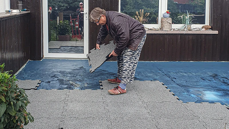 Se está renovando una terraza. Una mujer mayor coloca la siguiente baldosa de terraza de 18 mm de espesor con encaje en la terraza ya parcialmente cubierta con nuevas baldosas. Una lámina impermeabilizante sirve de base para el nuevo suelo de baldosas de goma. El encaje preciso de las baldosas permite una instalación fácil y estable.