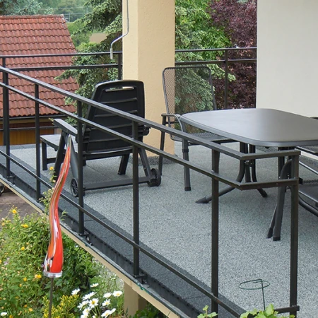 En la imagen se aprecia un balcón amplio, revestido con losetas WARCO de color gris. Una barandilla metálica negra rodea la zona y aporta seguridad. Una mesa espaciosa y sillas invitan a la convivencia. El piso elástico y antideslizante es resistente a la intemperie y fácil de mantener, brindando protección duradera y un espacio exterior cómodo para relajarse.