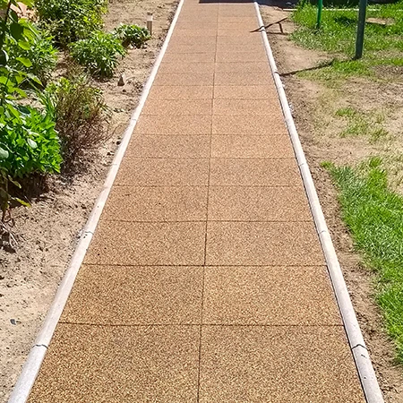 Un camino recto de jardín con baldosas cuadradas de goma y bordillo lateral atraviesa el parterre. Las baldosas marrones de granulado de caucho ofrecen una superficie antideslizante, resistente a la intemperie y de fácil mantenimiento. El camino asegura un paso limpio y seguro en todo clima y separa claramente los parterres del césped. Ideal para jardines, patios y accesos.