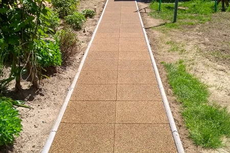 Un camino recto de jardín con baldosas cuadradas de goma y bordillo lateral atraviesa el parterre. Las baldosas marrones de granulado de caucho ofrecen una superficie antideslizante, resistente a la intemperie y de fácil mantenimiento. El camino asegura un paso limpio y seguro en todo clima y separa claramente los parterres del césped. Ideal para jardines, patios y accesos.